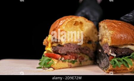 Schneiden Handwerk Burger ist Kochen auf schwarzem Hintergrund. Bestehen: sauce, Rucola, Tomaten, roten Zwiebeln Pommes, Gurken, Käse, Brötchen und Fleisch. Stockfoto