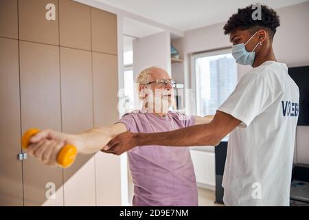 Ein älterer Bürger, der eine Kraftübung macht, wird von seinem Trainer unterstützt Stockfoto