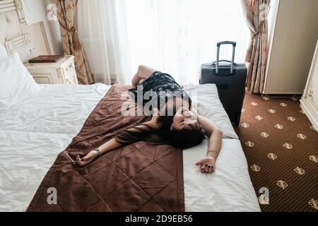 Glückliche junge Frau mit Gepäck kam in einem Hotelzimmer an. Erster Tag der Sommerferien. Auf dem Bett liegen. Fröhlicher Tourist in einer luxuriösen Unterkunft. Stockfoto