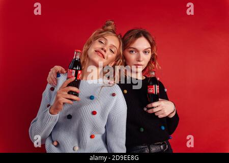Zwei hübsche junge Freundinnen halten Flaschen Cola. Ein Mädchen legte ihre Hand auf die Schulter eines anderen. Isoliert auf rotem Hintergrund. Stockfoto