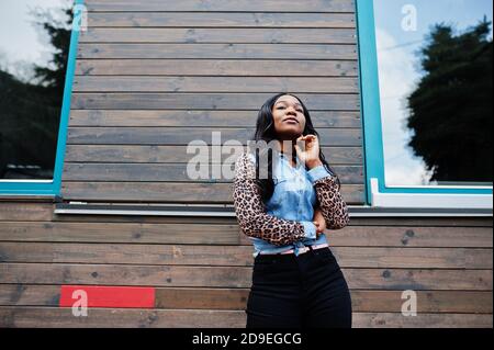 Hipster afroamerikanische Mädchen trägt Jeans Hemd mit Leoparden Ärmeln posiert auf der Straße gegen Holzhaus mit Fenstern. Stockfoto