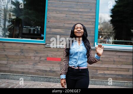 Hipster afroamerikanische Mädchen trägt Jeans Hemd mit Leoparden Ärmeln posiert auf der Straße gegen Holzhaus mit Fenstern. Stockfoto