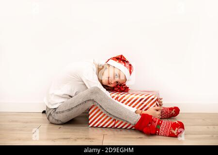 Ein schönes Mädchen in legerer Kleidung mit Weihnachtszubehör sitzt auf dem Boden und umarmt eine große Geschenkbox mit einem roten Band und einer Schleife. In voller Länge Stockfoto