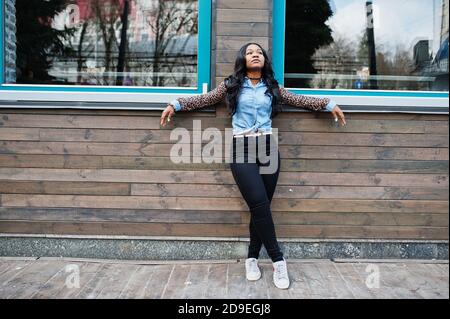 Hipster afroamerikanische Mädchen trägt Jeans Hemd mit Leoparden Ärmeln posiert auf der Straße gegen Holzhaus mit Fenstern. Stockfoto