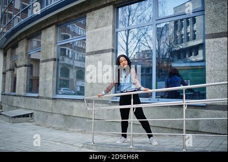 Hipster afroamerikanische Mädchen trägt Jeans-Shirt mit Leoparden Ärmeln posiert auf der Straße gegen moderne Bürogebäude mit blauen Fenstern. Stockfoto