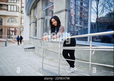 Hipster afroamerikanische Mädchen trägt Jeans-Shirt mit Leoparden Ärmeln posiert auf der Straße gegen moderne Bürogebäude mit blauen Fenstern. Stockfoto