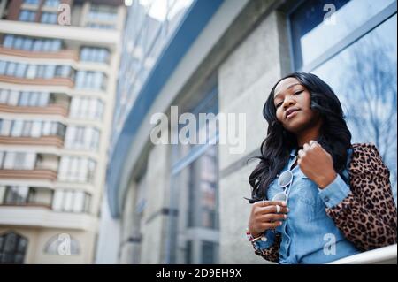 Hipster afroamerikanische Mädchen trägt Jeans-Shirt mit Leoparden Ärmeln posiert auf der Straße gegen moderne Bürogebäude mit blauen Fenstern. Stockfoto