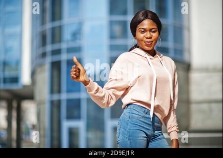 Hipster afroamerikanische Mädchen trägt rosa Hoodie, Jeans posiert auf der Straße gegen Bürogebäude mit blauen Fenstern. Stockfoto