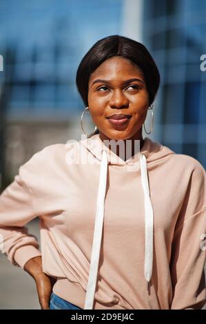 Hipster afroamerikanische Mädchen trägt rosa Hoodie, Jeans posiert auf der Straße gegen Bürogebäude mit blauen Fenstern. Stockfoto