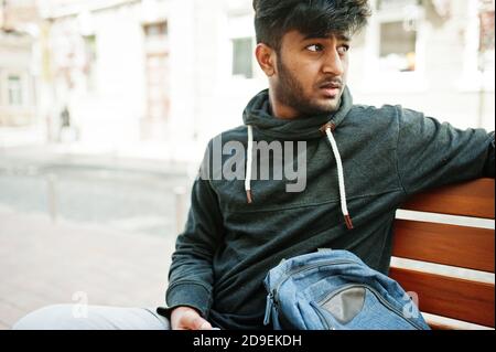 Porträt von jungen stilvollen indischen Mann Modell posieren in der Straße, sitzen auf der Bank und halten Smartphone zur Hand. Stockfoto