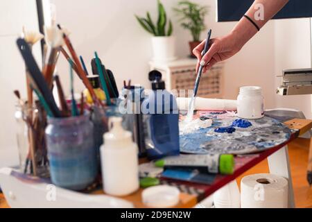 Die Hand der Malerin schmiert Farbe auf einer Palette in ihrem heimischen Atelier. Die Farbtöpfe und der Pinsel sind in Gläsern auf der Seite. Stockfoto