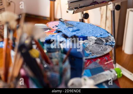 Die blaue Farbe des Malers schmierte sich im Atelier auf einer Palette. Die Farbtöpfe und der Pinsel sind in Gläsern auf der Seite. Stockfoto