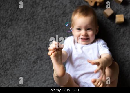 Nettes Baby spielt und liegt auf seinem Rücken auf einem Grauer Teppich mit Spielzeug Stockfoto