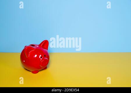 Rotes Schwein Sparschwein in einem weißen Fleck auf einem gelben Tisch vor dem Hintergrund einer blauen Wand. Leerzeichen für Text. Wohlstandskonzept. Speicherplatz kopieren Stockfoto