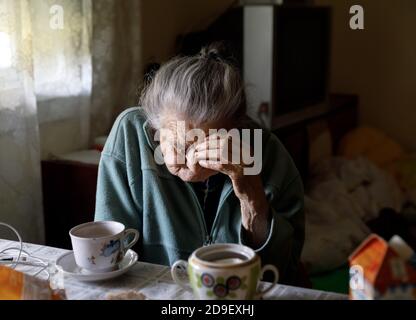 Alte depressive Frau. Eine ältere einsame Frau sitzt an einem Tisch in der Küche neben dem Fenster und trinkt Tee. Stockfoto