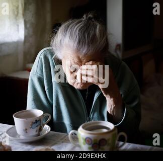 Alte depressive Frau. Eine ältere einsame Frau sitzt an einem Tisch in der Küche neben dem Fenster und trinkt Tee. Stockfoto