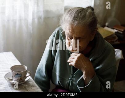 Alte depressive Frau. Eine ältere einsame Frau sitzt an einem Tisch in der Küche neben dem Fenster und trinkt Tee. Stockfoto