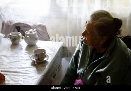 Alte depressive Frau. Eine ältere einsame Frau sitzt an einem Tisch in der Küche neben dem Fenster und trinkt Tee. Stockfoto