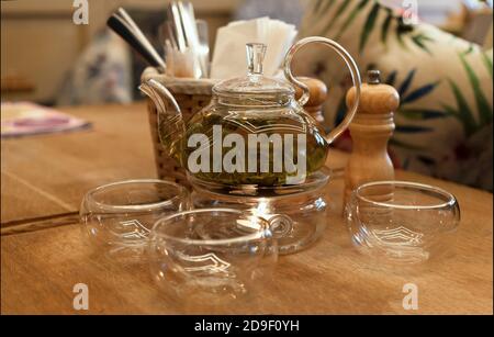 Transparente Teekanne mit grünem Tee und transparenten Tassen auf einem Holztisch in einem Restaurant. Stockfoto