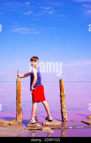 Der Junge lächelt. Gute Laune. Er hat ein weißgezahntes Lächeln. Ein Junge in roten Shorts auf einem Hintergrund von blauem Himmel und rosa See. Platz für Text kopieren. Stockfoto