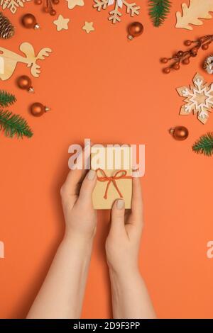 Womans Hände mit Pastell Maniküre halten Handwerk Geschenk oder Geschenkbox auf orange Pastell Tisch mit weihnachtsdekoration. Flache Lay-Ansicht von oben Stockfoto
