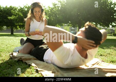 Junge kaukasische Paar trainiert Bauchmuskeln im Park - Mann tut ABS-Übungen mit seiner Freundin halten Zeit mit Eine Smartphone-App, die Stockfoto