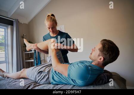 Junger Mann, der auf dem Krankenhausbett liegt und die Position der Gelenkschmerzen zeigt An die junge Frau Physiotherapeutin Stockfoto