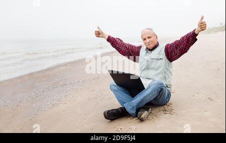 Kostenloses Lifestyle-Konzept. Glücklicher senor Geschäftsmann sitzt am Strand an einem nebligen Tag mit einem Laptop und genießt einen erfolgreichen Deal. Arbeiten während des Quarantänevorgangs Stockfoto