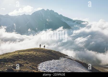 Nicht identifizierte Touristen mit großem Rucksack steht auf hohen Bergklippen Auf Höhe der Wolken vor dem Hintergrund der hohen schneebedeckten Bergkonzept Stockfoto