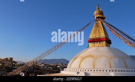 Riesige Boudhanath Stupa (Boudha) im Zentrum von Kathmandu, Nepal mit bunten buddhistischen Gebetsfahnen mit Gebäuden und Himalaya-Ausläufern geschmückt. Stockfoto