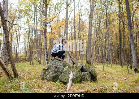 Kleiner Junge, der an einem Herbsttag auf einem großen Felsen im Wald klettert. Stockfoto