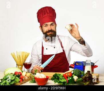 Kochen mit Lächeln in burgunderroten Uniform sitzt am Küchentisch mit Gemüse und Geschirr. Konzept der Restaurantküche. Der Küchenchef bereitet das Essen zu. Mann mit Bart hält Pilz und Messer auf weißem Hintergrund Stockfoto