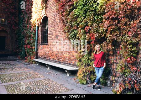 Junges Mädchen mit einem Spaziergang in der Herbststadt. Schöner Park mit trockenen gelben und roten Blättern und bunten Bäumen. Frau steht in der Nähe der Ziegelwand Stockfoto