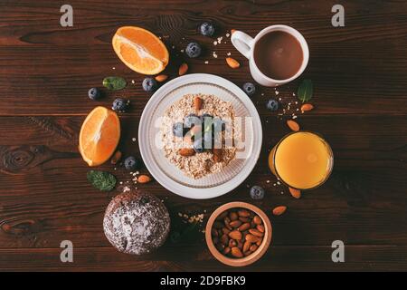 Gesundes Frühstück mit Haferflocken, Nüssen, Beeren, Orangen, Schokoladenmuffin und Tasse Kaffee auf braunem Holztisch, flach legen Zusammensetzung. Stockfoto