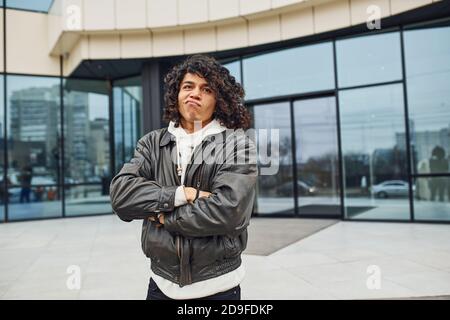 Schöner junger Mann mit lockigen schwarzen Haaren ist auf der Straße gegen Gebäude Stockfoto