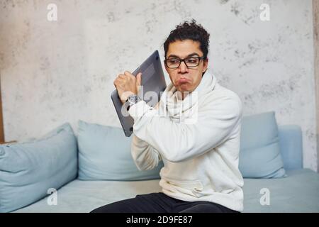 Wütend junger Mann mit lockigen schwarzen Haaren geht verrückt und Wütend auf Laptop in den Händen Stockfoto