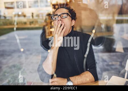 Junger Mann mit schwarzen lockigen Haaren sitzt drinnen im Café. Blick durch das Glas Stockfoto