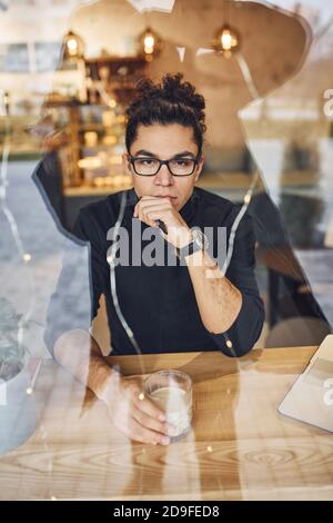 Junger Mann mit schwarzen lockigen Haaren sitzt drinnen im Café. Blick durch das Glas Stockfoto