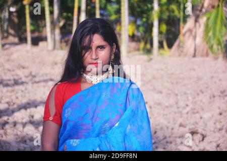 Nahaufnahme eines indischen bengalischen Teenager-Mädchen trägt blauen Sari Ohrringe Halskette rote Bluse mit langen dunklen Haaren in einem gepflügten Feld stehen. Stockfoto