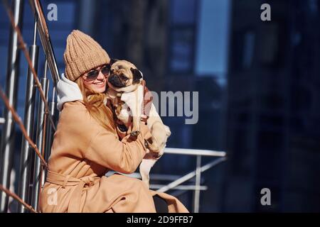 Frau in warmen Kleidern sitzen und haben Spaß mit ihr Kleiner Mops Hund in der Nähe des Geschäftsgebäudes, dass auf dem Hintergrund Stockfoto