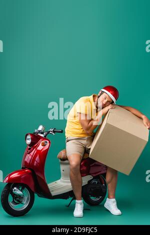 Müde Lieferung Mann im Helm hält großen Karton in der Nähe Roller auf blau Stockfoto