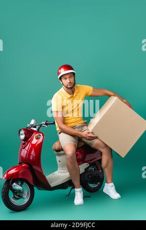 Erschöpft Lieferung Mann im Helm hält großen Karton in der Nähe Roller auf blau Stockfoto