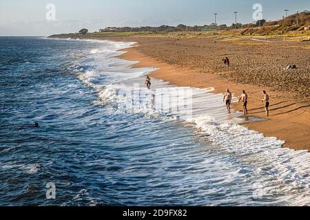 Alltag. Eine Gruppe von Männern beobachtete die Wellen am Strand an einem sonnigen, aber kalten Tag, während Damen zum Wasser kamen. Stockfoto