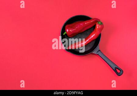 Zwei rote reife Chilischoten liegen auf einer kleinen schwarzen gusseisernen Pfanne auf einem leuchtend roten Hintergrund. Heiße Rabatte und Verkaufskonzept. Selektiver Fokus. Stockfoto