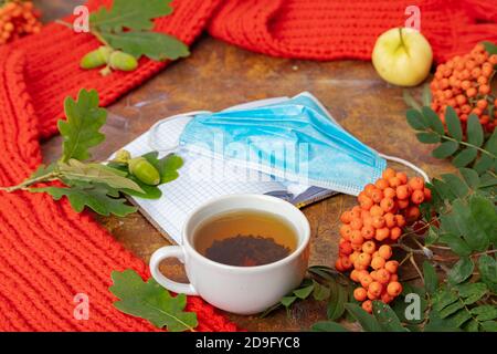 Eine medizinische Maske, ein roter Schal, eine Tasse Tee, eine Bergasche auf dem Tisch. Flatley. Konzept, Herbst, Quarantäne, covid. Hochwertige Fotos Stockfoto