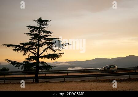 Dramatischer Sonnenuntergang über Wolken über einer Bergstraße. Auto auf der Seite in Sardegna, Italien geparkt. Silhouette eines Baumes. Stockfoto