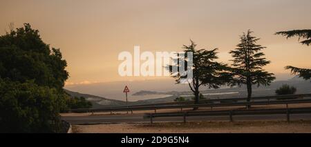 Gebogene Asphaltstraße und Biegeschild an einem heißen sonnigen Tag mit Mittelmeer hinter. Stockfoto