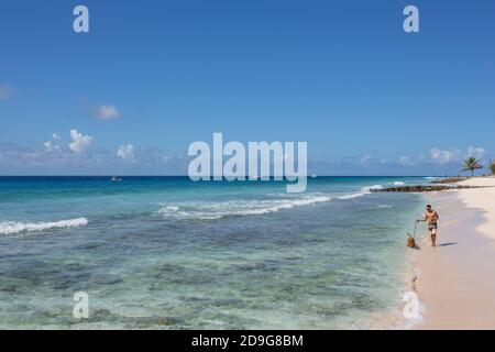 Ein attraktiver Mann spaziert mit dem Hund entlang des wunderschönen Sandstrandes in Barbados Stockfoto