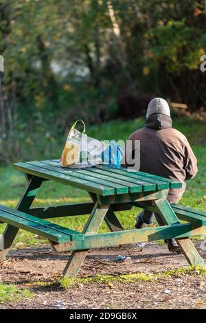 Ein Mann mit Hut sitzt allein an einer Parkbank und Tisch mit seinen Einkaufs-und Tragetaschen neben ihm auf dem Tisch. Stockfoto