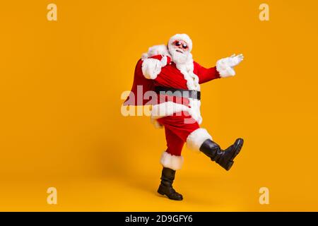 Ganzkörper-Profil Seitenfoto von santa claus mit groß Bauchbart tragen Weihnachtsgeschenk Geschenk Sack gehen tragen Kopfbedeckung Kappe Sonnenglas isoliert über hell Stockfoto